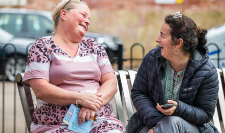 Two Individuals Engaged In A Conversation Sitting On A Bench Outdoors And Laughing