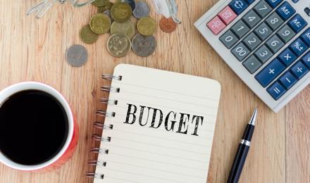 A notepad with budget written down alongside a hot drink, coins and a calculator