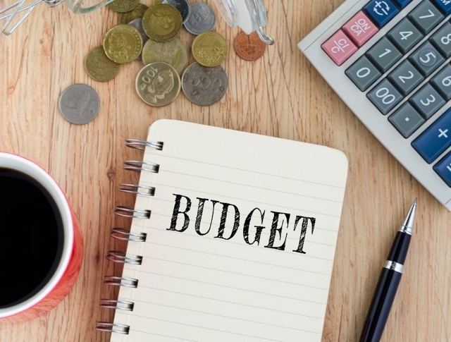 A notepad with budget written down alongside a hot drink, coins and a calculator